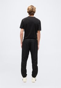 Νεαρό άτομο με κοντά ξανθά μαλλιά στέκεται γυρισμένο με την πλάτη, φοράει μαύρο μπλουζάκι, μαύρο φαρδύ παντελόνι και λευκά αθλητικά παπούτσια σε μονοχρωμο φόντο.