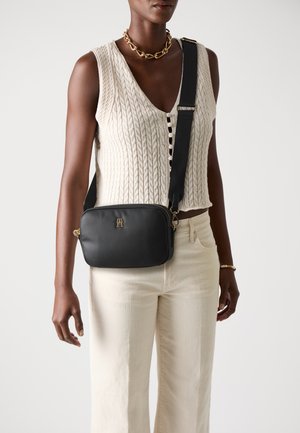 Bolso cruzado de cuero negro con un emblema dorado. Cuenta con cierre de cremallera, correa ajustable y textura suave. Llevado sobre una blusa de punto.