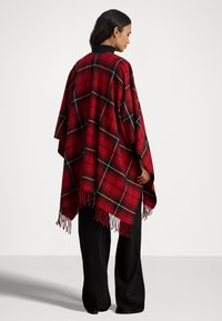 Poncho a quadri in rosso, nero e verde con bordi frangiati, realizzato in materiale morbido, presenta un colletto nero. Indossato sopra pantaloni larghi neri.