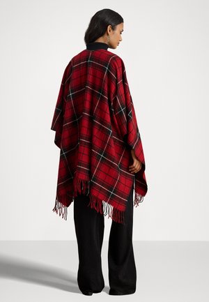 Poncho a quadri in rosso, nero e verde con bordi frangiati, realizzato in materiale morbido, presenta un colletto nero. Indossato sopra pantaloni larghi neri.