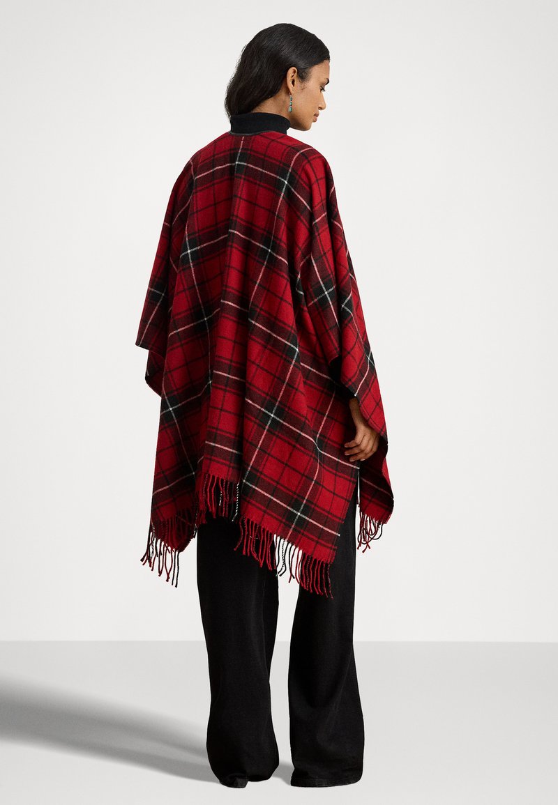 Poncho a quadri in rosso, nero e verde con bordi frangiati, realizzato in materiale morbido, presenta un colletto nero. Indossato sopra pantaloni larghi neri.