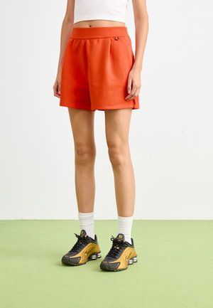 Pantalones cortos de algodón naranjas con cintura elástica y bolsillos laterales, combinados con zapatillas negras y doradas y calcetines blancos hasta el tobillo sobre un suelo verde.