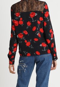 Blusa floreale con pannello in pizzo nero, con rose rosse su sfondo nero. Maniche lunghe e polsini con bottoni, abbinata a jeans blu strappati.