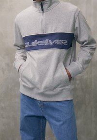 Persona vistiendo un sudadera gris con cremallera de cuarta parte y un panel azul, y un logo blanco de "Quiksilver", combinada con jeans azules, con las manos en los bolsillos de la sudadera.
