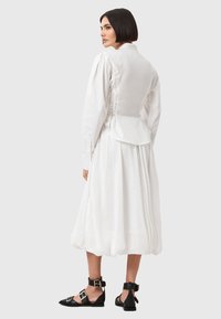 Biała sukienka z bufiastymi rękawami i regulowanym wiązaniem z tyłu. Posiada zwiewną spódnicę sięgającą do kolan, zestawioną z czarnymi sandałami na cienkich paskach.