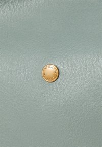 Superficie in pelle blu chiaro con una finitura texturizzata presenta un accento in metallo circolare dorato, impresso con la parola "COACH."