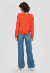 Maglietta rossa a maniche lunghe con righe testurizzate, abbinata a jeans a gamba larga di un blu chiaro e sneakers bianche. Vista posteriore, con una vestibilità comoda.