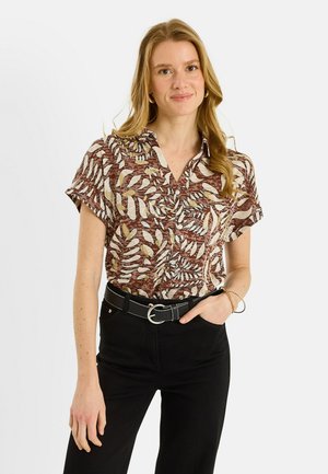 Mujer con camisa de manga corta de patrón de hojas en tonos marrón y beige, pantalones negros con cinturón, de pie con una mano en el bolsillo, sonriendo suavemente.