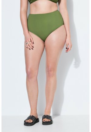 Frau trägt olivgrüne Bikinihose mit hoher Taille und schwarze Sandalen, steht vor weißem Hintergrund, Hände entspannt seitlich.