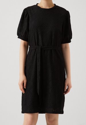 Musta polvipituinen mekko, jossa on tekstuurikuvio, pussihihat, pyöreä pääntie ja vyötärönauha, pukee henkilö, joka seisoo yksiväristä taustaa vasten.