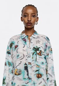 Camicia leggera, a maniche corte, multicolore con illustrazioni di elementi marini, fari e piante tropicali su uno sfondo bianco.