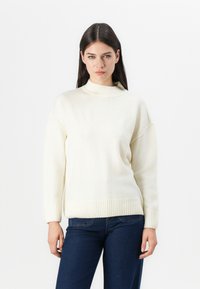 Hellbeige gestrickter Pullover mit hohem Kragen und geripptem Saum und Bündchen. Hat eine lockere Passform und eine weiche Textur. Wird mit blauen Jeans getragen.