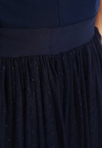 Abito navy con un corpino aderente caratterizzato da una texture liscia, una gonna a pieghe in tulle trasparente con pois e una cintura liscia a contrasto.