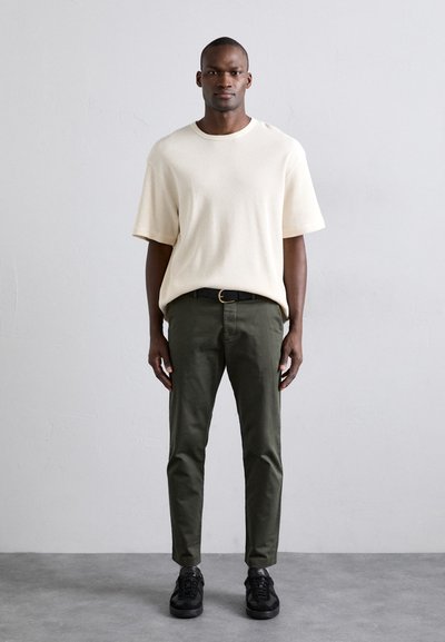 Cremfarbendes, strukturiertes Kurzarmhemd, olivgrüne Hose, schwarze Sneaker, vor einem neutralen Hintergrund stehend. Schlichtes Design, lässiges Outfit.