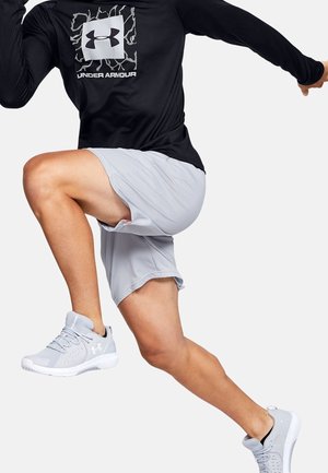 Person iført sort Under Armour langærmet trøje og lysegrå shorts midt i et hop, der viser atletiske sko og dynamisk bevægelse.