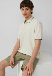 Hellbeige Poloshirt mit kurzen Ärmeln, grauen Kragenakzenten und einer Drei-Knopf-Leiste, kombiniert mit olivgrünen Shorts.