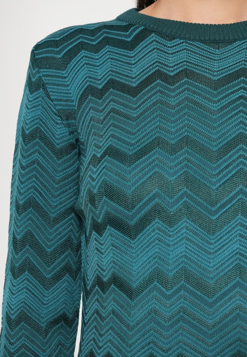 Pull en tricot teal avec un motif en zigzag, col rond et tissu texturé. Gros plan mettant en avant le design et la couleur.