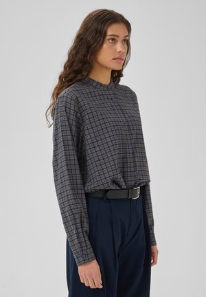 Vrouw met lang, golvend bruin haar draagt een blauw geruit overhemd ingestopt in een marineblauwe broek met een zwarte riem, staand tegen een effen achtergrond.