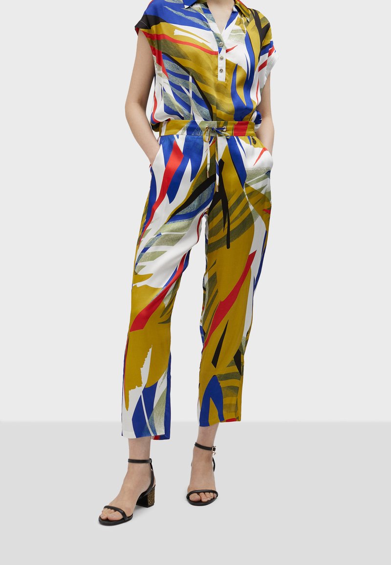 Tuta in satin con maniche corte, motivo astratto multicolore a foglie, vita cinchata e tasche laterali. Abbinata a décolleté neri con cinturini.