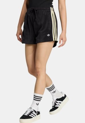 Persona che indossa pantaloncini Adidas neri con strisce bianche e gialle, calzini Adidas bianchi con strisce nere e scarpe da ginnastica Adidas nere con suole bianche.