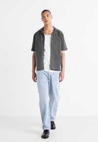 Camisa de manga corta, con botones, en tela gris oscuro, combinada con una camiseta blanca sin mangas, jeans de mezclilla azul claro y zapatos negros. Diseño y ajuste sencillos.