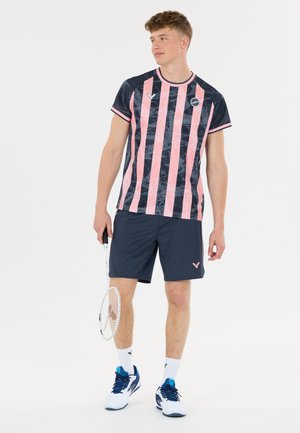 Ung mand holder en badmintonketcher, iført en pink og navystribet sportstrøje med navy shorts og hvide sportssko.
