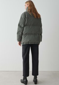 Giacca imbottita verde oliva con colletto alto, texture trapuntata e orlo elasticizzato, indossata sopra pantaloni dritti neri e stivaletti.