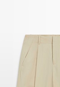 Beige high-waist pants with belt loops, front pleats, and slanted side pockets on a white background.