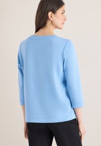 Femme aux longs cheveux foncés portant un haut bleu clair à manches trois-quarts et un pantalon noir, vue de dos sur un fond beige.