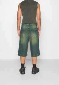 Pantalones cortos de mezclilla verde con un ajuste holgado, que cuentan con dos bolsillos traseros y un ligero desvanecimiento. Combinados con una camiseta de punto sin mangas de color oscuro y zapatillas negras.