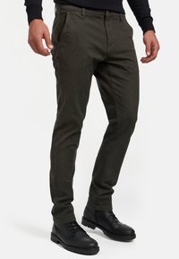 Pantalones ajustados de color verde oscuro, confeccionados en algodón texturizado. Presentan bolsillos frontales, un cierre de botón y detalles de costura sutiles. Combinados con botas negras.