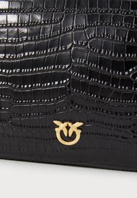 Mala de mão preta em couro com textura de crocodilo e um logotipo circular dourado com duas aves. Acabamento suave com detalhes de padrão em destaque.