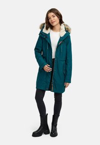 Giacca parka turchese con cappuccio foderato in pelliccia, dotata di zip frontale, coulisse regolabili e tasche laterali. Indossata sopra una maglietta bianca e jeans neri.