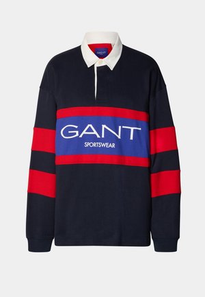 Camiseta de rugby azul marino de manga larga con rayas rojas en las mangas, cuello blanco y logo de "GANT SPORTSWEAR" en un panel azul y rojo en el pecho.
