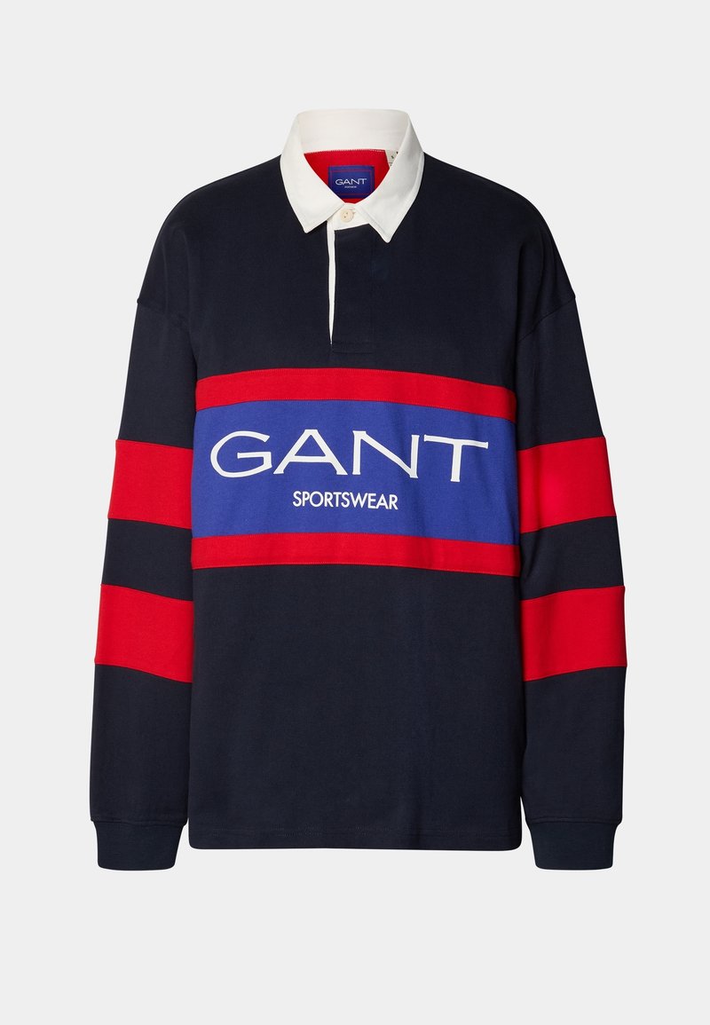 Maillot de rugby bleu marine à manches longues avec des rayures rouges sur les manches, un col blanc, et un logo "GANT SPORTSWEAR" sur un panneau bleu et rouge au niveau de la poitrine.