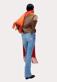 Suéter de punto marrón, jeans acampanados de un azul claro y una larga bufanda naranja con flecos. El modelo es visto desde atrás. Se pueden ver botas negras.