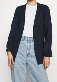 Granatowy dzianinowy kardigan z otwartym przodem, d�ługimi rękawami i prążkowaną fakturą. W zestawie z jasnoniebieskimi jeansami z wysokim stanem oraz białą koszulką.