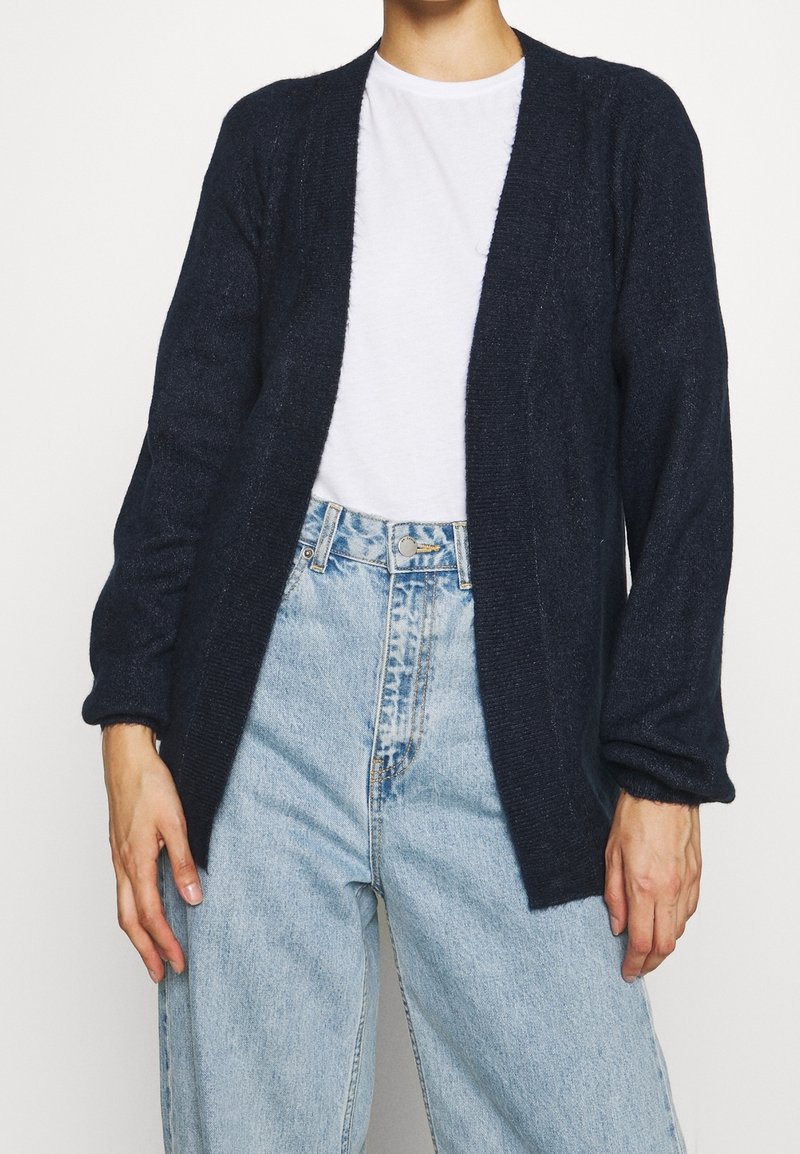 Granatowy dzianinowy kardigan z otwartym przodem, długimi rękawami i prążkowaną fakturą. W zestawie z jasnoniebieskimi jeansami z wysokim stanem oraz białą koszulką.