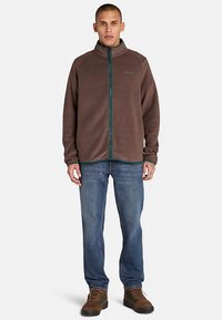 Chaqueta de forro polar marrón con cuello alto y cremallera verde, usada sobre vaqueros azules y botas de senderismo marrones. Diseño sencillo y funcional.