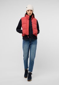 Rode puffervest over een donkerblauwe zip-up fleece, gecombineerd met blauwe jeans en navy schoenen. Kenmerkt een geribbelde beige beanie.