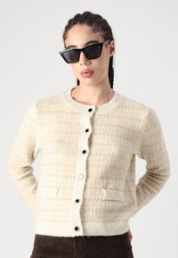 Gilet texturé beige avec des accents crème, doté d'un col rond, d'une fermeture à boutons et de deux poches avant. Des boutons noirs rehaussent le design.