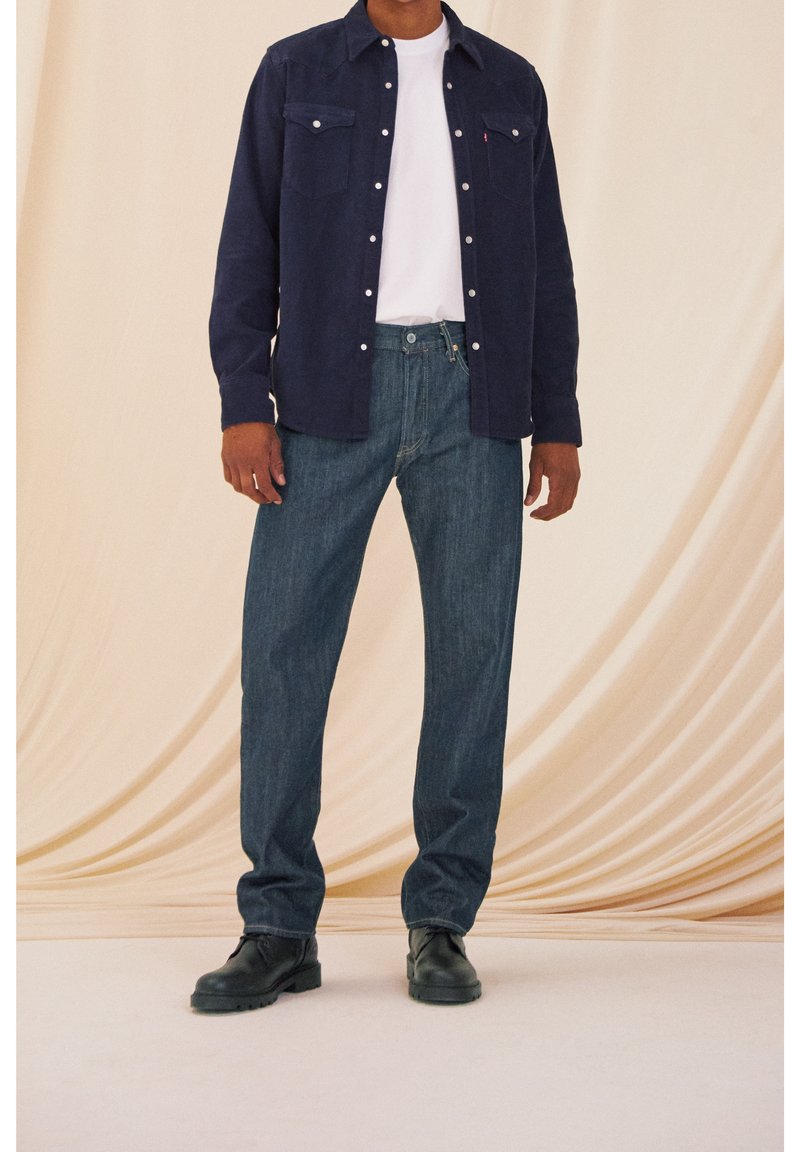 Homme portant une chemise bleu foncé à boutons par-dessus un t-shirt blanc, un jean bleu et des bottes noires, debout devant un fond beige clair drapé.