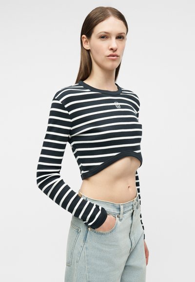 Νεαρή γυναίκα που φοράει ένα μαύρο και λευκό ριγέ cropped τοπ με μακριά μανίκια και ανοιχτόχρωμο μπλε τζιν, με το ένα χέρι στην τσέπη της.