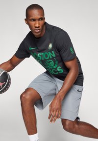 Czarna koszulka Nike z zielonym napisem i logo "BOSTON CELTICS", w połączeniu z szarymi szortami do koszykówki. Gracz trzyma piłkę do koszykówki z marką Jordana.