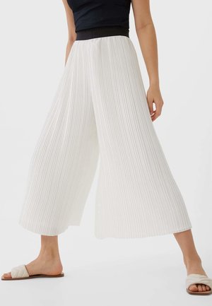 Femme portant un pantalon blanc à jambes larges plissé, un haut sans manches noir et des sandales plates blanches matelassées, debout devant un fond clair uni.