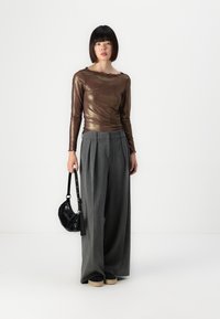 Bronze, long-sleeve top with a textured pattern, paired with wide-leg, pleated grey trousers. Holding a small, glossy black bag.