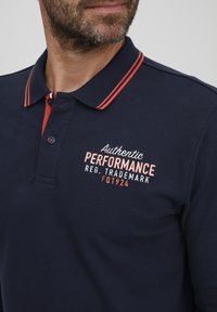 Námornícka polo košeľa vyrobená z bavlny s červeno-bielym prúžkovaným golierom. Obsahuje vyšívaný text "Authentic Performance" v červenej a bielej farbe.