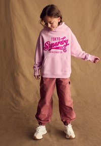 Felpa con cappuccio rosa con un grande logo, abbinata a pantaloni cargo rosa larghi e scarpe da ginnastica bianche. Il tessuto sembra morbido e confortevole.