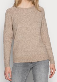 Beige stickad tröja med långa raglanärmar, rund halsringning och ribbat avslut. Mjuk textur med ett subtilt melerat mönster.