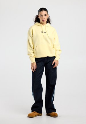 Jonge vrouw staat en draagt een lichtgeel "New York" hoodie, losse donkere spijkerbroek met lichtblauwe strepen en bruine werkschoenen op een witte achtergrond.